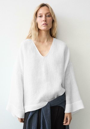 Žena s dlouhými blond vlasy, která má na sobě volný texturovaný bílý top s výstřihem do V a tmavý svetr uvázaný kolem pasu, na jednobarevném pozadí.