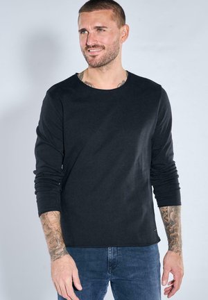 Lächelnder Mann mit kurzen Haaren und Tätowierungen trägt ein schwarzes Langarmshirt und blaue Jeans, steht vor einem schlichten hellen Hintergrund.