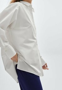 Chemise blanche à manches longues avec un col classique, présentant une coupe ample, des boutons et un ourlet arrondi, associée à un pantalon bleu marine.