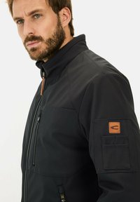 Sort softshell jakke med høj krave, lynlås fortil og et struktureret stof. Indeholdende en brystlomme og et brunt læderlogo-patch påærmet.