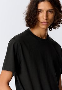 Černé bavlněné tričko s krátkými rukávy, kulatým výstřihem, měkkou texturou a volným střihem. Omezené detaily zdůrazňující jednoduchost.