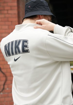 Personne portant une veste blanche Nike avec un grand logo dans le dos et un chapeau bob noir, ajustant le col à l'extérieur près d'un mur en briques.