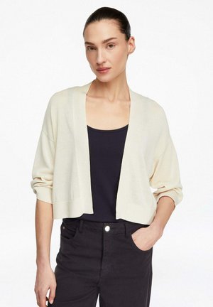 Kobieta ubrana w kremowy krótki sweter na guziki na czarny top na ramiączkach i czarne dżinsy z wysokim stanem, pozująca z jedną ręką w kieszeni.