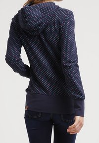 Fleece hoodie med marin bakgrund och flerfärgade prickar. Har en huva och enfärgade marinmanschetter och nederkant.