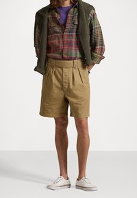 Person med rutigt skjorta över lila t-shirt, kaki veckade shorts, vita sneakers och en olivgrön tröja över axlarna.