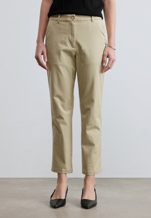 Kvinne iført beige bukser med rette ben, svarte spisse hæler, en svart topp og et sølvarmbånd, stående på et grått gulv.
