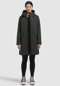 Mörkgrön, lång vattentät kappa med huva, frontknappar, sidofickor och justerbar dragsko. Tillsammans med svarta leggings och skor.