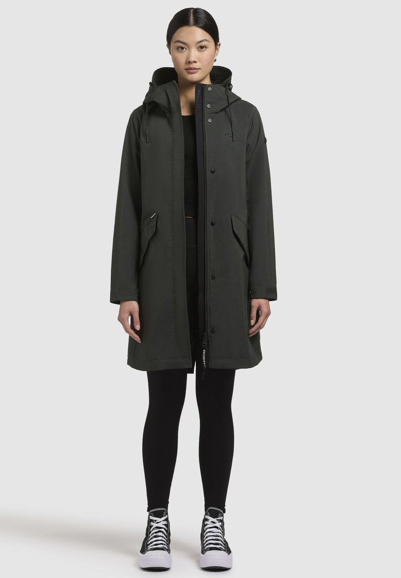Mörkgrön, lång vattentät kappa med huva, frontknappar, sidofickor och justerbar dragsko. Tillsammans med svarta leggings och skor.