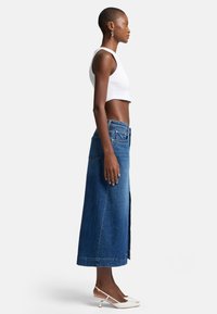 Jupe maxi en denim bleu moyen avec une haute fente, associée à un débardeur court blanc et des chaussures à talons ouverts blanches. Texture lisse et design décontracté.