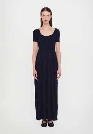 Maxi dress in navy, ribbed upper body with a scoop neckline, flowing pleated skirt, short sleeves, paired with black flat shoes.
