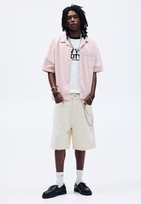 Camicia rosa chiaro a maniche corte con bottoni sopra una t-shirt bianca con grafica, abbinata a pantaloni corti bianco sporco e scarpe nere, con dettagli in catena argentata.