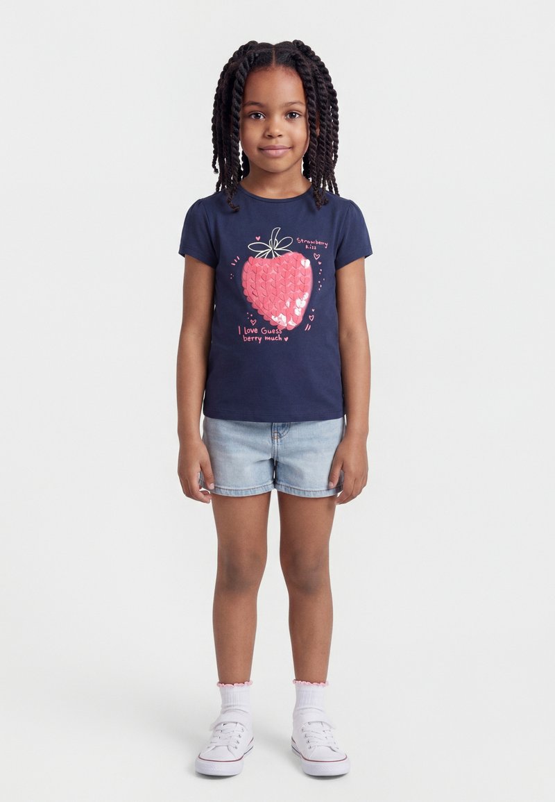 Νεαρό κορίτσι με πλεξούδες, φορώντας μπλε σκούρο μπλουζάκι με ροζ σχέδιο φράουλας και ανοιχτόχρωμα κοντά τζιν σορτς, στέκεται με τα χέρια χαλαρά.