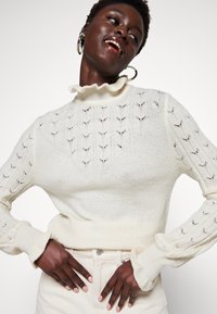 Maglione a maglia color crema con un colletto alto arricciato, delicati motivi a foglia sulle maniche e polsini voluminose. Indossato con pantaloni di colore chiaro.