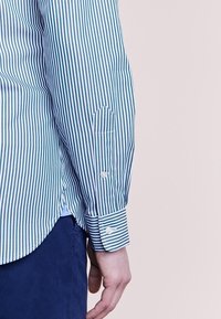 Randig skjorta i grön och vit bomull, med knappad manschett, sidofente och spetsig fickdetalj på ärmen.