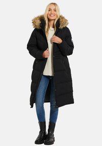 Sort lang pufferjakke med en hætte med pelskant, frontlynlås, sidelommer og quiltet design, båret over en cremet sweater og blå jeans.
