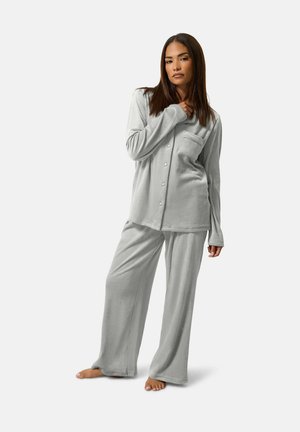 Femme debout pieds nus portant un haut à manches longues boutonné en tissu doux gris clair et un pantalon large de pyjama sur fond blanc.