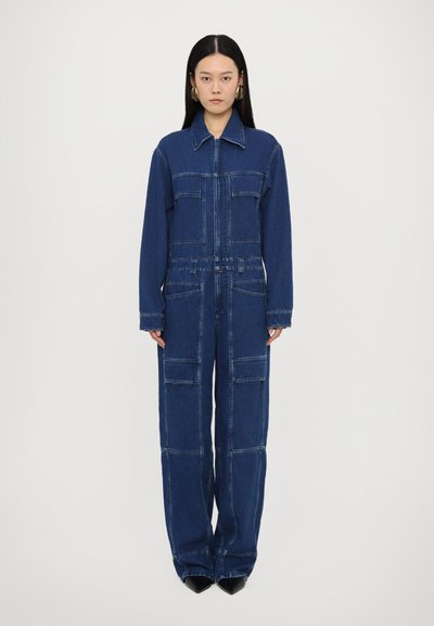 Denim jumpsuit i mørkeblå med lange ærmer, frontlynlås, flere lommer og kontrastsyning; bred benlængde og afslappet pasform.