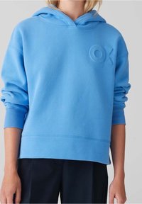 Osoba v jasně modré mikině s kapucí, s vyhrnutými rukávy a vyraženým logem "OX" na hrudi, v černých kalhotách.