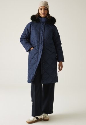 Kobieta ubrana w długi granatowy pikowany płaszcz z czarnym futrzanym kołnierzem, beżową dzianinową czapkę, granatowe spodnie z białym paskiem i białe trampki.