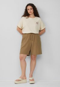 Beige T-shirt met korte mouwen en een zonnekleurige graphic, gecombineerd met bruine textuurshorts met een elastische tailleband en trekkoord. Lichtgewicht plateausandalen.