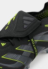 Black football boot with textured upper, green accents, and gray detailing. Features a distinctive velcro flap and a clear sole.