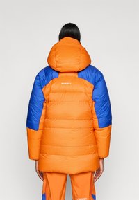 Jachetă puffer cu un corp portocaliu și mâneci albastre, cu un model texturat în grilă și o glugă mare. Logo-ul este afișat pe spate.