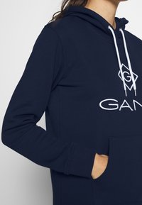 Marineblauwe hoodie met een voorzak, witte trekkoorden en een logo op de borst. Gemaakt van zacht materiaal, met ribbelmanchetten.