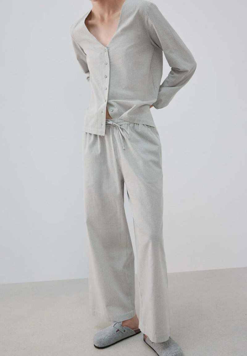 Personne portant un haut boutonné à manches longues gris clair et un pantalon large avec cordon, assorti à des chaussons gris à enfiler sur un sol clair.
