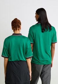 Camisetas polo verdes de manga curta com uma textura subtil, apresentando um colarinho preto e detalhes brancos. Duas pessoas vistas de costas.
