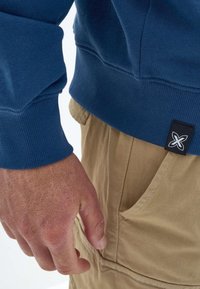 Felpa blu navy con polsini e orlo a costa, con un'etichetta del logo nera. Pantaloni beige con una tasca laterale visibile accanto alla mano.