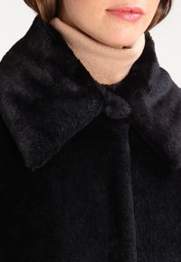 Czarny sztuczny futrzany płaszcz z dużym kołnierzem i zapięciem na guziki, charakteryzujący się gładką, miękką teksturą. Pod spodem widoczny jest jasnobeżowy sweter z golfem.