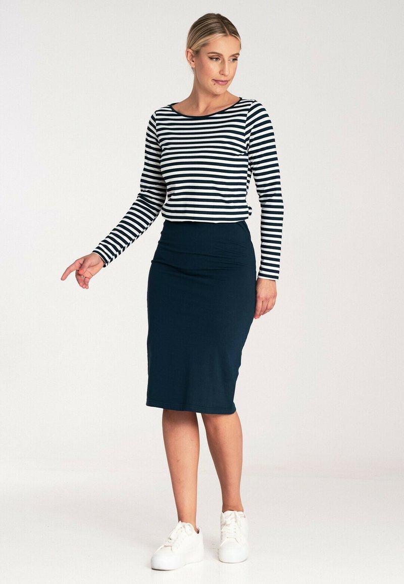 Pasiasta koszula z długim rękawem w granatowo-białe paski, zestawiona z dopasowaną granatową spódnicą ołówkową i białymi trampkami, stojąca na jednolitym tle.