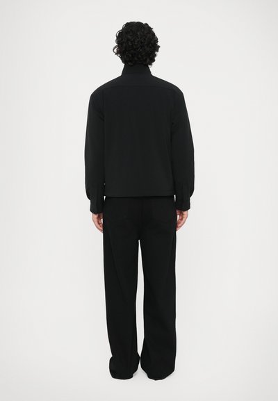 Muž s kučeravými vlasmi, oblečený v čiernej bunde a voľných čiernych nohaviciach, stojaci chrbtom k jednoliatej bielej pozadí.