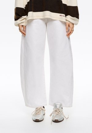 Pantaloni a gamba larga bianchi in denim con una texture liscia. Indossati con sneakers grigio chiaro dotate di dettagli riflettenti.