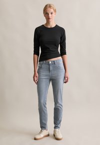 Vrouw staat tegen een effen achtergrond, draagt een zwart longsleeve shirt, lichtblauwe jeans en beige sneakers, kijkt recht vooruit.