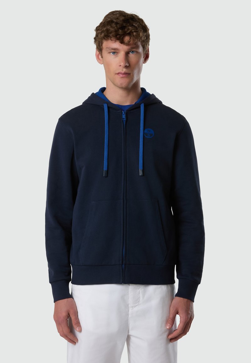 Joven con sudadera azul marino con cremallera, cordones azules y un pequeño logo en el pecho, combinado con pantalones blancos, de pie frente a un fondo gris.