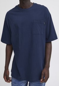 Tricou din bumbac bleu marin, cu guler rotund, mâneci scurte și un buzunar pe piept. Prezintă o croială dreaptă și un design minimalist.