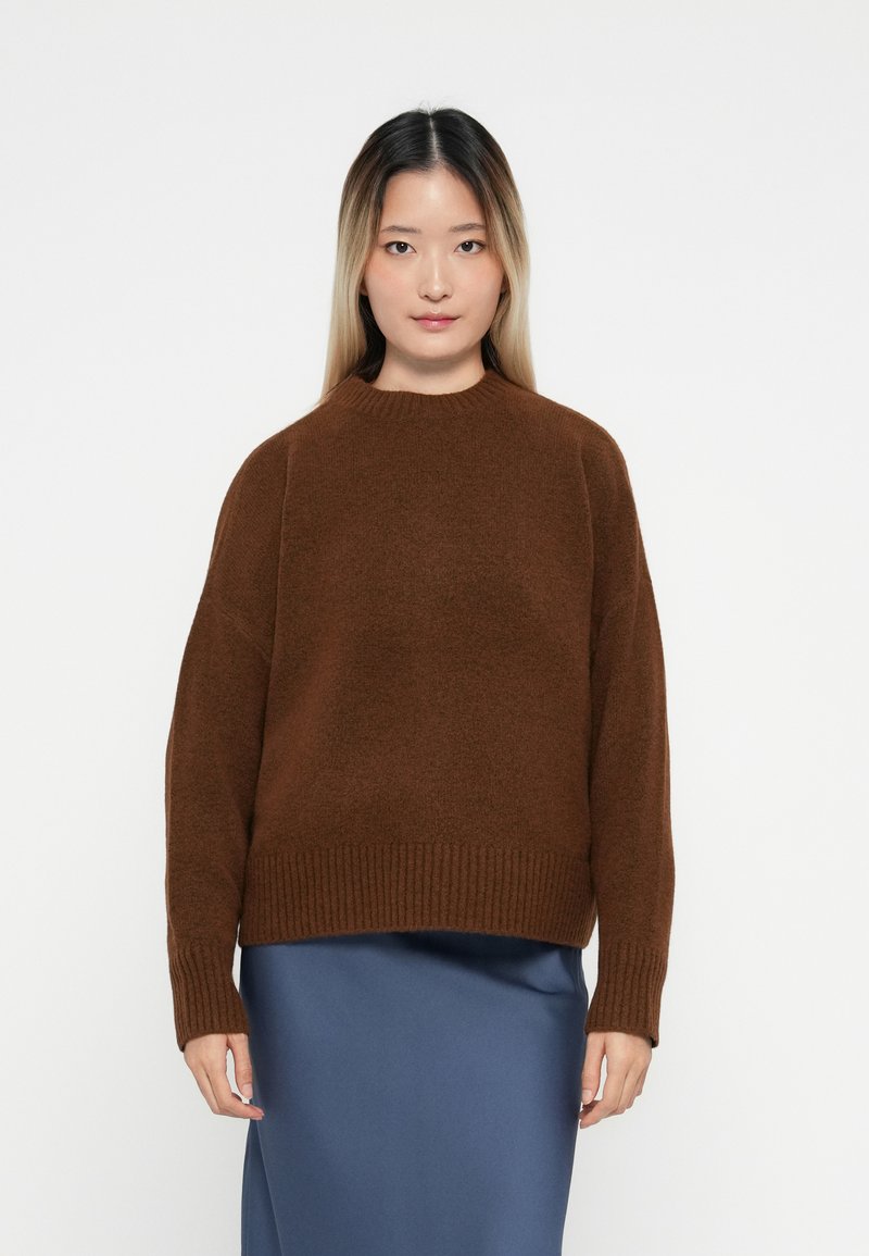 Žena s rovnými, dlouhými ombre vlasy, oblečená v hnědém oversized pleteném svetru a modré saténové sukni, stojící na bílém pozadí.