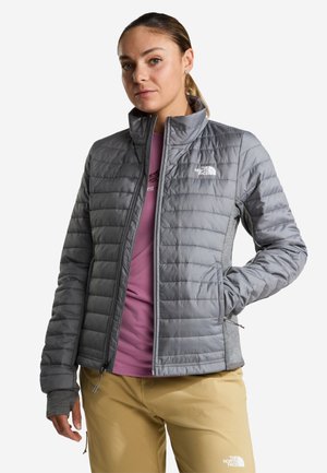 Donna che indossa una giacca trapuntata grigia, una camicia rosa e pantaloni beige con i loghi The North Face, in piedi con una mano in tasca.