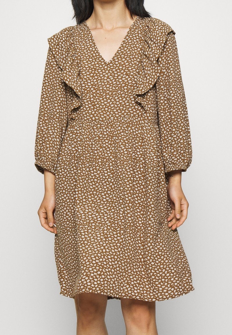 Femme portant une robe fleurie marron avec un col en V volanté et des manches longues, les mains détendues le long du corps sur un fond uni.