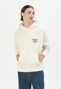 Felpa beige realizzata in tessuto morbido, con tasca frontale e cappuccio con cordino. Mostra un logo blu navy sulla parte sinistra del petto.