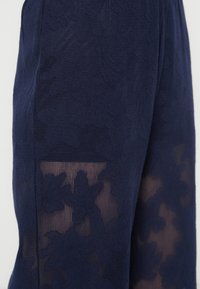 Μπλε ναυτικό παντελόνι με πάνελ από φλοράλ δαντέλα με υφή σε επιφάνεια. Το ύφασμα είναι ημιδιαφανές, αναδεικνύοντας περίτεχνα σχέδια.