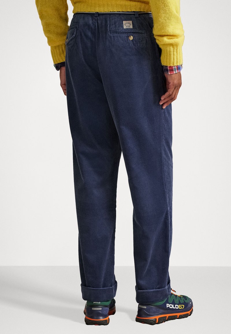 Marineblaue Cord-Hosen mit lockerer Passform, ausgestattet mit einer Gesäßtasche mit Logo-Patch und umgeschlagenen Säumen, kombiniert mit bunten Sportschuhen.