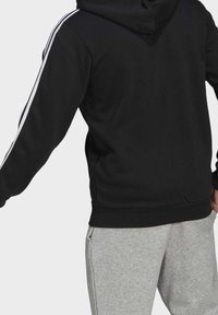 Sweat à capuche noir avec des bandes latérales blanches, doté d'une capuche à cordon de serrage et de poignets côtelés. Associé à un pantalon de survêtement gris clair. Vue arrière uniquement.