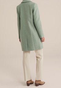 Pitkä mintunvihreä villakangastakki, jossa on jäsennelty muotoilu, lovikaulus ja suora siluetti. Yhdistetty kermaisiin housuihin ja ruskeisiin korkokenkiin.