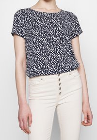 Námořnická modrá krátkosrstá blůza s bílými puntíky; kulatý výstřih; spárovaná se světlými džíny s knoflíkovým zapínáním.