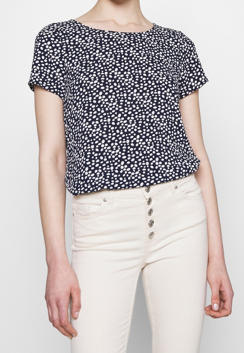 Námořnická modrá krátkosrstá blůza s bílými puntíky; kulatý výstřih; spárovaná se světlými džíny s knoflíkovým zapínáním.