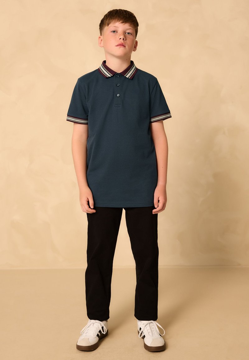 Türkisfarbenes Poloshirt mit gestreiftem Kragen in Burgunderrot, Creme und Blau; kombiniert mit schwarzen Hosen und weißen Turnschuhen. Kurzärmlig, lässige Passform.