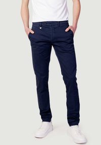 Pantaloni chino blu navy in cotone, con zip, tasche laterali e design slim fit, abbinati a sneakers bianche.