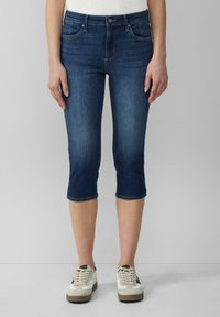 Tmavomodré denim capri nohavice so priliehavým strihom, stredne vysokým pásom, dvoma prednými vreckami a manžetami na koncoch. Nosené s bielymi teniskami.